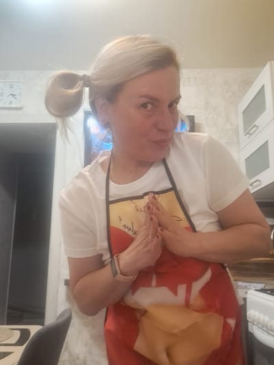 A woman with blonde hair tied in a ponytail, wearing a white t-shirt and an apron with a maple leaf design, is standing indoors. She is facing the camera with her hands clasped in front of her. The background includes kitchen cabinets, a television, a clock, and a dining area with a chair. There is no verifiable evidence to confirm the location is Elektrostal, Russia.