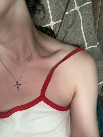 The image shows a close-up of a person's upper body, focusing on their neck, shoulder, and upper chest. They are wearing a white tank top with red trim and a silver cross necklace. The person has dark hair that is partially visible. The background consists of a patterned fabric, possibly bedding. There is no discernible indication of the location within the city of Postoyata, Ukraine.