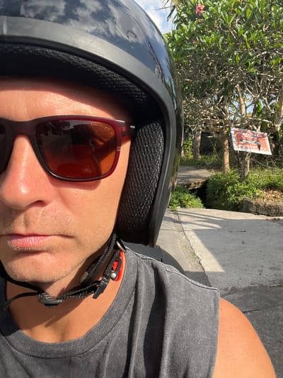 A selfie taken from the perspective of a person wearing a black helmet and sunglasses, seated on a vehicle in Sukawati, Indonesia. The person's face, from the sunglasses down to the neck, is visible, along with a portion of their grey t-shirt. The background features lush green foliage, trees, a paved path, and a sign. The sign appears to be a warning, with text that includes "PERINGATAN GARAGE" and a phone number. The sky is visible at the top of the image, reflecting on the helmet. The lighting suggests it is daytime.