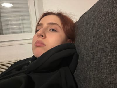 An upside-down view shows a woman with fair skin, auburn hair, and wearing a black hoodie. The hoodie's hood partially covers her face. Her eyes are visible, and she looks directly at the camera. The background includes a gray, textured surface, possibly a couch, and a white wall with a window covered by horizontal blinds. The light from the window illuminates part of her face.