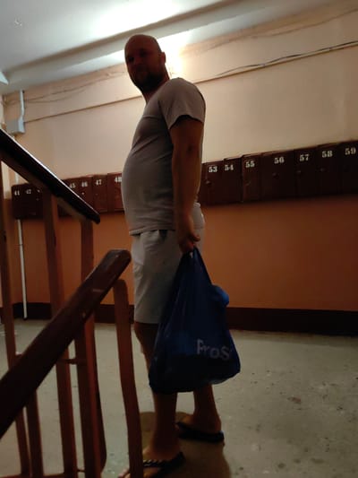 A man is walking up a staircase in an apartment building, carrying a blue shopping bag. He is wearing a light grey t-shirt and light grey shorts. The staircase has a wooden railing. On the wall of the stairwell, there are a row of mailboxes with numbers ranging from 41 to 59. The man appears to be bald with a beard and is looking over his shoulder. The lighting suggests it is daytime. The overall impression is a mundane, everyday scene in a residential building. The blue bag has the word "Pros" visible on it.