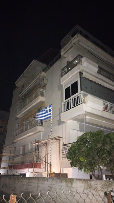 The image shows a multi-story building at night in Perea, Greece. A Greek flag is prominently displayed on a flagpole in front of the building, extending from approximately the second to the fourth floor. The building appears to be made of concrete and has several balconies with white railings and awnings. Scaffolding is visible on the lower levels of the building. A tree with green foliage is located to the right of the building, and a concrete wall with a wire fence runs along the bottom of the image. The scene is illuminated by artificial light, casting shadows and highlighting the textures of the building and its surroundings. The sky is dark, indicating nighttime.