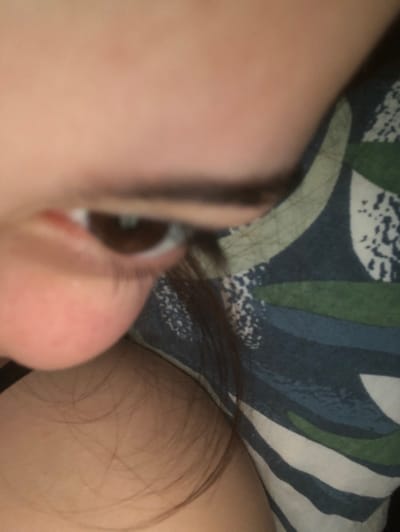 An extreme close-up captures a person's face from a distorted, low-angle perspective. The eye, with long eyelashes and a visible reflection, dominates the right side, adjacent to a pink nose. The rest of the face is soft and out of focus. Strands of dark hair frame the right side of the face, resting against pale skin. A patterned blue and green fabric serves as the backdrop.