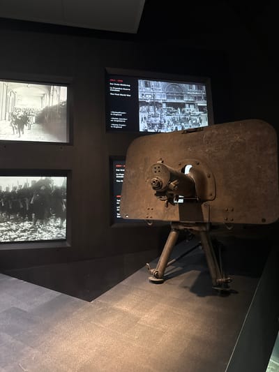 This media file depicts a museum exhibit related to World War I, likely located in Bern, Switzerland. The central focus is a large, weathered metal object, possibly a piece of military equipment or artillery, mounted on a stand. The object is positioned diagonally across the frame, obscuring parts of the background displays.
To the left of the equipment, two screens are visible. The upper screen displays a black and white historical photograph, likely depicting a scene from the war, with many people gathered. Below this, another screen shows text in multiple languages, including German, French, and English, with dates and titles such as "1914-1918," "Der Erste Weltkrieg," "La Première Guerre mondiale," and "The First World War." Further text indicates themes like "Heimatfront" and "Home Front."
Below these screens, two more screens are partially visible. The left one shows another black and white historical photograph, seemingly of people in procession or gathered in a formal setting with grand architecture. The right screen displays a black and white image of a large group of people, possibly soldiers or a crowd, in what appears to be an outdoor setting. The overall lighting is dim, typical of a museum environment, highlighting the displays. The setting appears to be an indoor exhibition hall. The images and text suggest a historical exhibition focusing on the impact of World War I.