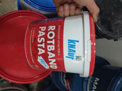 In Arinj, Armenia, a close-up shot focuses on a red bucket with a white and red plastic container of "ROT BAND PASTA" held by a hand. The container is decorated with Russian text and the "KNAUF" logo. The hand, belonging to an individual whose fingers are visible, is gripping the container. The background reveals other buckets, some blue and some black, suggesting a storage or workspace environment. The overall scene is indoors, with the lighting illuminating the primary subject.