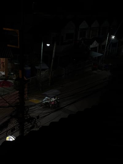 This is a nighttime, high-angle, outdoor shot of a dimly lit urban scene in Kathu, Thailand. The image is dominated by darkness, with scattered areas of light revealing details. The primary subjects are the complex network of electrical wires and cables that hang precariously above a street or alleyway.
On the left side of the frame, a multi-story building is partially visible. It appears to be weathered, with corrugated metal roofing and some exposed structural elements. A bright, circular light fixture is visible on this building, casting a strong beam downwards. Another building element on the left has a semi-circular light, possibly the moon.
The right side of the image shows a sloped, paved surface, likely a street or sidewalk, also cluttered with numerous wires. Some laundry or fabrics are draped over railings or structures, appearing as colorful blurs in the dim light. One section of the building on the left has what looks like a metal staircase or fire escape.
In the foreground, a dark, horizontal structure, possibly a balcony or a crossbar, is visible, further obscuring the scene below and emphasizing the tangled mass of wires. Another building element with a yellow, U-shaped marking is visible in the lower left quadrant. In the far right, a circular object with a yellow and white pattern is illuminated.
The time of day is clearly night, given the darkness and artificial lighting. The weather is not discernible. The location cues, such as the building styles and the dense wiring, are characteristic of some urban areas in Southeast Asia. There are no discernible emotions, as the scene is devoid of clearly visible human figures engaged in any specific activity. No text is legible in the image.