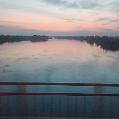 The image shows a wide river at dusk, with the sky displaying soft hues of pink, orange, and blue. The water reflects the colors of the sky, creating a serene and tranquil scene. Silhouetted trees line the distant banks of the river, and a faint mist hangs over the water in the background. In the foreground, two dark horizontal railings are visible, suggesting the viewpoint is from a bridge or elevated structure. The overall atmosphere is peaceful and contemplative, with a sense of calm as day transitions into night.
