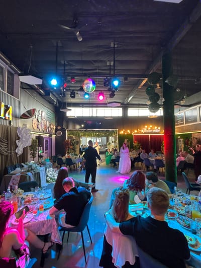 The image depicts a festive indoor gathering, possibly a wedding reception or a party, held in Chernushka, Russia. The scene is illuminated by a variety of colorful lights, including blue, pink, and disco ball effects, creating a vibrant atmosphere. Several round tables are set up, adorned with white tablecloths, floral arrangements, and food platters, indicating a meal is in progress.
In the foreground, several individuals are seated at a table, engaged in conversation and looking at their phones. One person is holding a smartphone, illuminated by its screen. In the middle ground, a man in a suit is standing, seemingly addressing a group of people. A woman in a long white dress, likely the bride, is positioned near the center of the room, facing away from the camera. A line of guests are seated at tables in the background, observing the events.
The environment appears to be a banquet hall or a restaurant with industrial-style walls and exposed ceilings. Decorative elements include a large green column covered in artificial foliage and a wall adorned with numerous clocks. The overall impression is one of a lively and celebratory occasion.