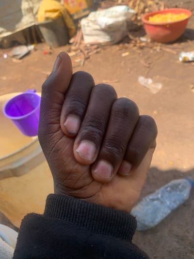 A close-up, first-person perspective shot shows a clenched fist with dark skin and trimmed fingernails, partially obscuring the background. The hand is covered by the sleeve of a black sweatshirt. In the blurred background, a purple plastic cup sits within a larger light-colored bucket. Further back, a red bowl filled with yellow kernels, possibly corn, is visible, alongside a clear plastic water bottle lying on the ground. The ground appears to be dirt with scattered debris. The scene suggests an outdoor environment, possibly in Buga da Gindi, Nigeria.