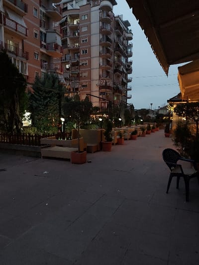 This image captures a quiet urban scene in Tiranë, Albania, during dusk or early evening. The sky is a soft, muted grey-blue, indicating twilight, and artificial lights are beginning to illuminate the surroundings.
On the left, a tall, multi-story apartment building stands prominently. It has a light-colored facade with reddish-brown accents and features numerous balconies, some with distinctive curved shapes and others rectangular, all adorned with red railings. Many windows are visible across its many floors, with some interior lights already turned on.
To the right, a wide, paved pedestrian path or plaza extends from the foreground into the distance. Along this path, a series of square planters, alternating in color between reddish-brown and lighter tones, hold small trees or shrubs. Several glowing lampposts are spaced along this planted border, casting a warm light. On the paved surface in the middle-right, a dark, possibly plastic, outdoor chair is visible.
Trees and other foliage are interspersed between the apartment building and the pathway. In the upper left corner, a dark awning or roof overhang suggests the photograph was taken from a covered outdoor area, possibly a cafe or balcony. No people are present, and there is no visible text. The scene evokes a calm and peaceful atmosphere as the city transitions from day to night.