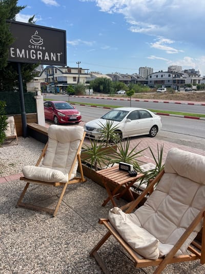 The image depicts an outdoor seating area of a cafe, possibly the "Emigrant Coffee" as indicated by a sign. The seating area is paved with small, multi-colored pebbles and features two wooden deck chairs with light-colored cushions, a small wooden table, and several potted plants. In the background, a street is visible with cars parked and driving, including a red Hyundai sedan with a Turkish license plate "34 GE 4772" and a white sedan. Across the street, there are buildings with modern architecture and some greenery. The sky is partly cloudy, suggesting a daytime scene in a city. The overall impression is of a relaxed outdoor cafe environment, likely located in a contemporary urban setting in Antalya, Turkey, given the Turkish license plate.