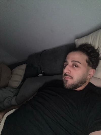 A man with dark hair in a bun, a beard, and a black t-shirt reclines on a gray couch. He rests his head on a pillow, looking directly at the camera. A phone charger is visible near the couch. Several pillows and a blanket are bundled near the man's waist. The background features a gray wall.