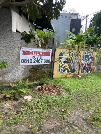 The image shows a gray concrete wall with a red and white sign attached to it. The sign advertises the sale of a property by Discovery Property with the contact number 0812 2467 600. The sign also reads "Dijual Rosie" which translates to "For Sale Rosie". The wall is adjacent to a rusty yellow metal gate with graffiti on it. The ground is covered in green grass and brown leaves. The scene is located in Lembang, Indonesia, at the coordinates -6.834 latitude and 107.603 longitude.