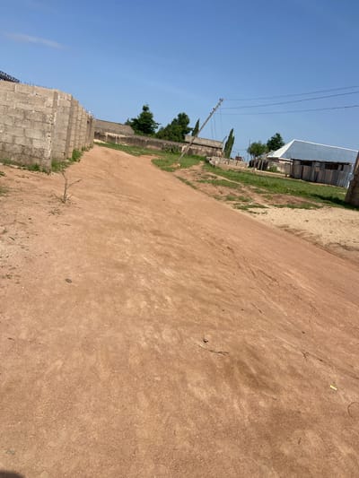 The image depicts an unpaved dirt road in Kuson, Nigeria, under a clear blue sky. To the left, a partially constructed wall made of concrete blocks runs along the road. Further down the road, trees are visible, and to the right, a building with a corrugated metal roof and siding stands adjacent to the road. A wooden utility pole with wires is also present in the background. The road surface is reddish-brown and appears dry and dusty. A few sparse, dry twigs are scattered on the road. The overall scene suggests a rural or semi-urban environment with ongoing construction and basic infrastructure.