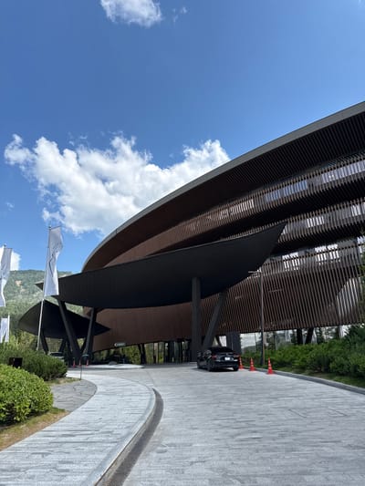 The image shows a modern architectural building with a curved, dark gray roof and a slatted wooden facade. The building has large windows that reflect the sky. In front of the building, there is a paved walkway with a textured, light gray surface. A black car is parked on the right side of the walkway, and three orange traffic cones are placed in a line behind it. Lush green bushes and trees are visible in the background, with a hint of a mountainous landscape. The sky is a vibrant blue with scattered white clouds. Two white flags with dark text are planted on the left side of the walkway, partially obscured by the building's overhang. One flag is higher than the other. The scene appears to be an entrance or arrival area to a contemporary establishment, possibly a hotel or resort, given the flags and the grand architecture. The lighting suggests it is daytime, with bright sunlight. There is no discernible activity or people engaged in any specific event. The overall impression is one of modern design and natural surroundings.