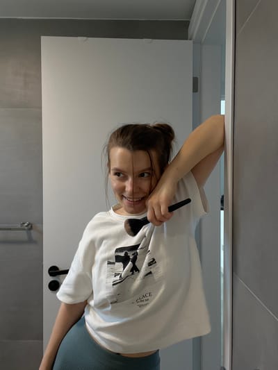 A young woman with her hair in a bun, wearing a white t-shirt and teal leggings, is leaning against a doorway and holding a makeup brush. She is smiling and looking to her left. The background appears to be a modern bathroom with gray tiled walls and a white door. The lighting suggests an indoor setting, possibly a home or hotel. This scene is not identifiable as being in Limassol, Cyprus.
