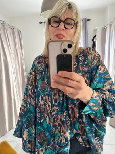 An indoor mirror selfie taken in Chambœuf, France, depicts an individual from the chest up. The person, with blonde hair and wearing dark, round-rimmed glasses, is shown sticking their tongue out while holding a light-colored smartphone with a dual-camera system. They are wearing a loose-fitting, long-sleeved blouse featuring a multi-colored abstract and paisley pattern in shades of teal, blue, brown, red, and beige, along with dark lower garments. A silver-toned ring is visible on the ring finger of the hand holding the phone. The background, reflected in the mirror, consists of a white wall, a grey bowl-shaped ceiling light fixture, and light-beige curtains on metal rods on both sides. A white tiled floor is visible at the bottom, alongside a partial view of a yellow upholstered object in the lower left and a white shelving unit with dark clothing on the right.