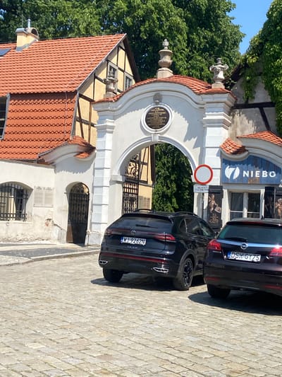 The image depicts an exterior scene in the city of Świdnica, Poland, focusing on a historic arched gateway. The gateway is constructed of white stone with ornate architectural elements, including decorative urns at the top and a circular inscription plaque above the arch. The roof of the adjacent building is covered in red tiles.
In the foreground, two cars are parked on a cobblestone street. The car on the left is a black Volkswagen Taigo with the license plate WJ-9262N. The car on the right is a dark-colored Opel Insignia with the license plate DSW GY75. The cars obscure a portion of the gateway and the street.
Behind the gateway, lush green trees are visible, indicating an outdoor setting. To the right of the gateway, a sign with the number "7" and the word "NIEBO" (Polish for "sky" or "heaven") is mounted on a blue background, suggesting a business or establishment. Another circular sign with a red border and a white circle within it, a common traffic sign indicating a prohibition, is also present.
The overall environment appears to be a public space, possibly a historical district or a courtyard, given the architectural style and the cobblestone paving. The image is taken on a clear, sunny day, as indicated by the bright lighting and the clear blue sky visible through the trees.