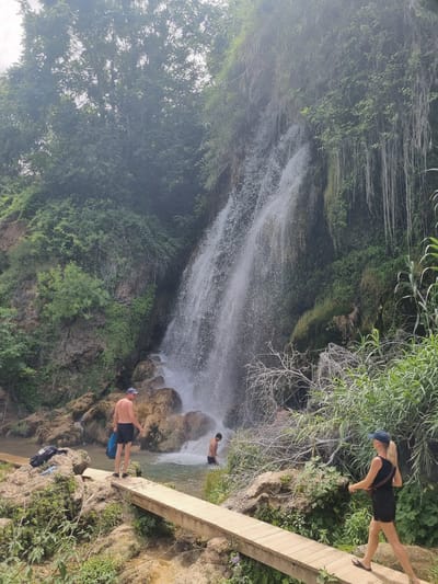 A group of people are at a waterfall in the city of Anna, Spain. In the foreground, a woman with blonde hair, wearing a blue cap, black tank top, and black shorts, walks on a wooden bridge. To her left, a man with dark hair, wearing a blue cap and black shorts, stands on a rock near the waterfall. Further left, a man with dark hair and a blue cap, wearing black shorts, stands on a wooden walkway. In the background, a waterfall cascades down a rocky cliff face covered in greenery. The scene appears to be a natural park or recreational area.
