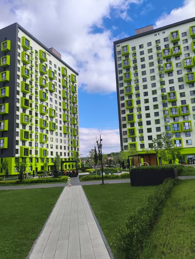 The image depicts a modern residential complex in Penza, Russia, under a partly cloudy sky. Two tall, multi-story apartment buildings with a striking architectural design dominate the scene. The buildings feature a light-colored facade accented with numerous lime-green, protruding window frames, giving them a contemporary and vibrant appearance. The building on the left has a section of its facade painted dark gray.

In the foreground, a paved walkway extends through a well-maintained grassy area. A young child on a scooter is visible on the walkway, appearing to be playing or exploring the open space. Behind the child, a group of people are seated under a covered structure, possibly a gazebo or seating area within the complex. Trees and manicured hedges are strategically placed throughout the complex, adding to the landscape. The time of day appears to be afternoon, with bright daylight and clear blue sky peeking through the clouds. The overall impression is one of a modern, family-friendly living environment. No visible text is present in the image.