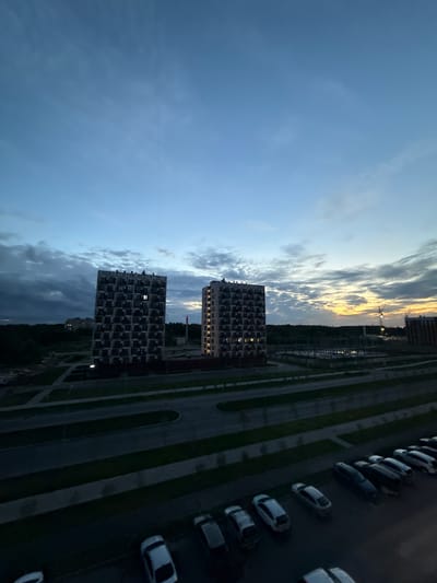 The image depicts a twilight scene in a developing urban area, likely a residential district in Veliky Novgorod, Russia. Two prominent, multi-story apartment buildings with dark, checkered facades stand in the mid-ground. The building on the left has a single lit window. In front of the buildings, there is a paved area that appears to be a street or parking lot, bordered by green strips of grass or hedges. Further back, the landscape transitions to a dark treeline, and beyond that, a construction site with a visible crane and unfinished structures suggests ongoing development. The sky above is a blend of deep blue and lighter shades of gray and yellow, indicating either the end of sunset or the beginning of dawn, with wispy clouds present. In the foreground, a row of parked cars is visible on a dark surface, most of which are light-colored sedans and SUVs. The overall impression is of a quiet, modern residential area undergoing expansion under a dramatic sky.