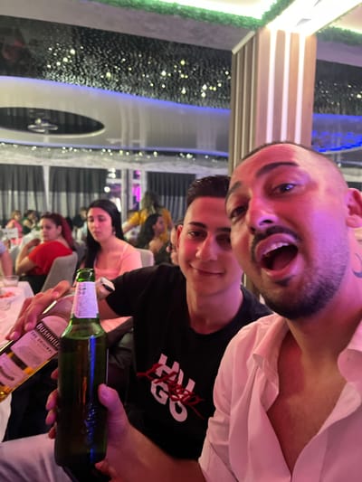 The image depicts an indoor social gathering in Sevlievo, Bulgaria. In the immediate foreground, two men are visible. The man on the right, with dark hair, a beard, and a crescent moon tattoo on the left side of his neck, is wearing a white collared shirt; his mouth is open in mid-expression. He is holding a green glass bottle in his left hand. To his left, a younger man with dark hair, wearing a black t-shirt, is smiling slightly and holding a clear glass bottle labeled "Bushmills The Original Irish Whiskey" in his right hand.
The background shows multiple individuals, predominantly women, seated at tables, indicating a restaurant or event hall setting. Notable figures include a woman in a red top and another in a light peach-colored top. The environment features a dark ceiling adorned with numerous small, bright lights, creating a starry effect. Blue and purple accent lighting strips are visible along the ceiling and walls. Vertical, illuminated light-colored columns and dark curtains are present in the mid-ground. Green decorative foliage lines portions of the ceiling structure. The overall scene is illuminated by artificial light, with a prominent pinkish-purple ambient hue. White tablecloths are visible on the tables.