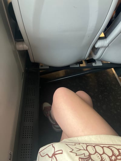 The image shows a close-up view of a person's legs, wearing a light-colored shirt with a graphic print of grapes and a pair of sneakers. The person is seated, with their legs crossed. The setting appears to be indoors, possibly on a public transport vehicle given the presence of a white, curved structure that resembles a toilet or a seat on a train or bus. The flooring is dark and speckled, and a metallic vent with a grid pattern is visible. The lighting is neutral, suggesting an indoor environment without strong natural light. No specific time of day or weather conditions can be inferred. The location context of Krakow, Poland is provided, but there are no explicit visual cues within the image to confirm this. The overall mood is candid and everyday, with no strong emotions conveyed. There is no visible text in the image.