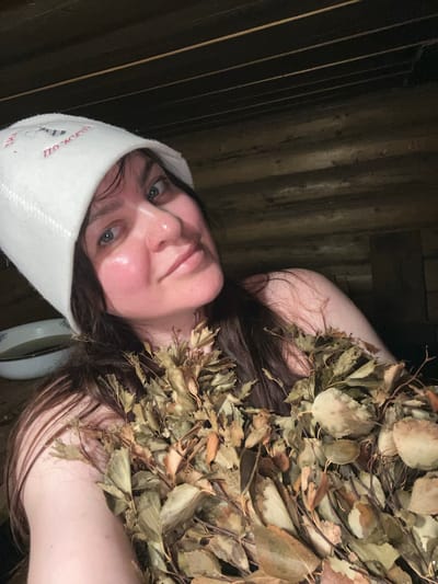 A woman is lying down, wearing a white sauna hat with text and a logo, and covered in dried leaves. The background appears to be wood paneling, possibly inside a sauna. A round object resembling a bowl is partially visible near her head. The location is purported to be in Protasy, Russia, though this cannot be independently verified from the image alone.