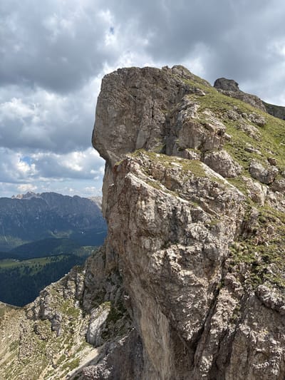 The image shows a dramatic, mountainous landscape from a high vantage point. The foreground is dominated by a large, weathered rock formation, casting a shadow and creating a sense of depth and scale. The rock face is textured and appears rugged, with patches of green grass growing in crevices. Below the rock overhang, a vast valley unfolds, filled with rolling green hills and dark, forested slopes. In the distance, more rugged mountain peaks can be seen, some with snow-capped summits. The sky is a mix of dramatic, dark clouds and patches of brighter sky, suggesting variable weather, possibly a storm brewing or passing. The lighting is somewhat diffused, but the sun is still strong enough to highlight the textures of the rock and the distant landscape. The overall mood is one of awe and perhaps a touch of trepidation due to the steep drop and the imposing nature of the rock formations. There are no people or specific objects visible in the frame, making it a purely natural landscape shot. The orientation of the image is vertical, emphasizing the height and the vastness of the scene. The location appears to be a high alpine area, likely in a region known for its dramatic mountain scenery.