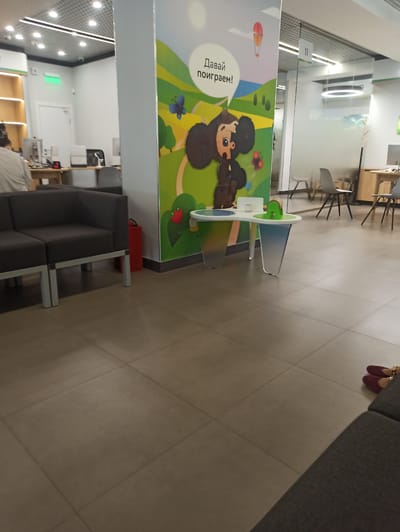 The image depicts the interior of a modern office space or waiting area, likely in Atabe, Georgia. The floor is tiled with large, light-brown square tiles. A dark grey sectional sofa is positioned in the foreground on the left. To the right of the sofa, a large, colorful mural featuring a cartoon character with large ears and a speech bubble with Russian text is prominent. Next to the mural, a white and gradient colored table with circular indentations and a green toy is situated. In the background, glass partitions and modern office furniture, including chairs and desks with computers, are visible. The lighting appears to be overhead fluorescent and circular recessed lighting. A red fire extinguisher is mounted on the wall near the sofa. The overall impression is of a clean, well-lit, and contemporary commercial environment. There are no visible people in the immediate foreground or engaged in specific actions, though shadows suggest movement or presence just outside the frame. The environment suggests a place designed for customer service or as a waiting area.