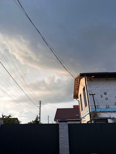 The image captures a cloudy sky with some shafts of sunlight breaking through, suggesting either early morning or late afternoon. The scene is dominated by the sky and the upper parts of buildings, including a structure under construction with white brickwork and exposed wooden beams. Another building with a dark tiled roof and a smaller window is partially visible. A tall, dark metal fence runs along the right side of the frame. Several utility poles with numerous wires crisscross the sky, indicating an urban or suburban setting. The overall impression is one of an overcast sky with dramatic lighting. There is no visible text in the image. The orientation of the image is rotated 90 degrees counter-clockwise.