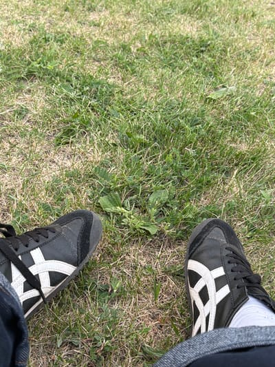 The image shows a person's legs, wearing black athletic shoes with white stripes, sitting on grass. The shoes appear to be Onitsuka Tiger sneakers. The grass is a mix of green and dry blades. The person is wearing dark blue jeans. The scene is outdoors, suggesting a park or open grassy area in Naberezhnye Chelny, Russia. The lighting indicates it is daytime.