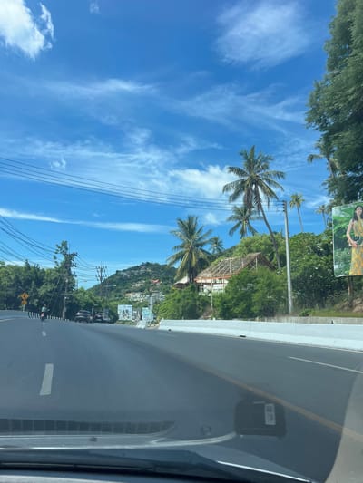 The image depicts a street scene in Baan Chaweng Noi, Thailand, under a bright blue sky with scattered white clouds. The road curves to the left, with a white line marking the lane. A motorcycle is visible in the distance on the left side of the road, along with several cars. Dense green foliage and palm trees line the roadside, with a hillside dotted with houses in the background. A large billboard featuring a woman in a yellow dress is prominent on the right side of the road. Power lines stretch across the sky, supported by utility poles. The scene appears to be captured from the perspective of a moving vehicle.