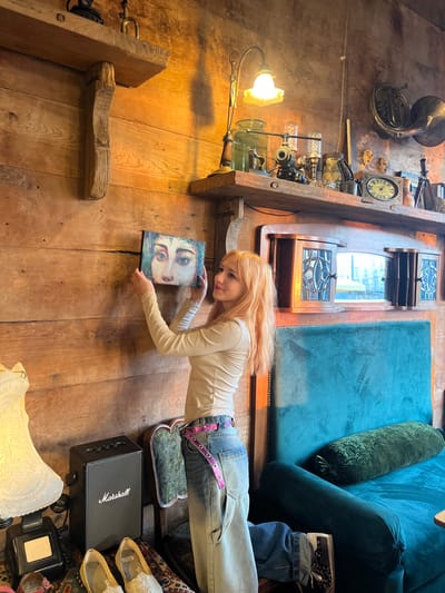The image captures an indoor scene, appearing to be a cozy, eclectic, or rustic-themed room, shot from an elevated perspective looking down. The main subject is a young woman with long, flowing, light orange or blonde hair, smiling softly and looking towards the camera. She is lying on a textured wooden floor, extending her body diagonally across the frame.
She is dressed in a simple, long-sleeved, light cream or off-white top and light blue, loose-fitting jeans. A striking detail is her shiny, metallic purple belt adorned with numerous eyelets, cinching her waist. In her hands, she holds a small, rectangular canvas or picture frame featuring an abstract painting of eyes or a stylized face with prominent, colorful eyes.
The room is richly detailed with various antique and decorative items. To the left of the woman, on what appears to be a wall or a sturdy wooden shelf, rests an antique French horn, a vintage-style clock with roman numerals, and an array of glass bottles and intricate metallic fixtures, possibly part of a unique lighting arrangement or a decorative display resembling old scientific equipment. A warm, yellow-toned light emanates from a nearby lamp with a glass shade, casting a soft glow on the wooden surfaces. Heavy wooden beams and structures are also visible, contributing to the rustic ambiance.
In the upper right portion of the image, a large, plush teal or dark blue velvet sofa or seating area is visible, accompanied by a dark green velvet bolster pillow, suggesting a comfortable lounging space. Near the woman's feet, a pair of light-colored, possibly glittering or patterned, flat shoes are placed on a patterned rug or textile. A black Marshall brand speaker is positioned nearby, confirming its presence with the visible "Marshall" logo. Further into the background, a wooden cabinet or display case with glass panes reflects some light, adding to the room's character. Another decorative white object, possibly a lamp base or vase, is partially visible in the bottom right corner. The overall impression is one of warmth, character, and artistic sensibility.