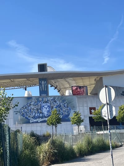 The image captures an exterior view of the Estádio do Dragão in Porto, Portugal, on a clear day. The stadium's facade features a large mural depicting a group of football players in blue and white jerseys, with the text "CAMPEÃO" and "Conseguimos" visible. A digital screen on the stadium displays a close-up of a person's face wearing glasses. A large banner with a football jersey is suspended from the stadium's roof. In the foreground, a chain-link fence with overgrown grass and foliage separates the street from the stadium. Traffic signs and trees are also present.