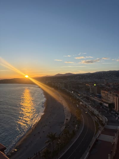 This high-angle shot captures the Baie des Anges in Nice, France, during sunset. The low-lying sun on the western horizon emanates intense golden rays across the sky and reflects brightly on the dark blue-green waters of the Mediterranean Sea, where white-capped waves break on the shore. A wide, curved pebble beach is populated by numerous small, indistinct human figures. Parallel to the beach, the Promenade des Anglais features a wide pedestrian pathway and an adjacent multi-lane asphalt road with visible white lane markings and scattered vehicles. Palm trees line sections of the promenade. Inland, a dense urban area of multi-story buildings with varied facades extends towards a backdrop of dark, silhouetted hills and mountains. The sky transitions from clear blue overhead to warm orange and yellow hues near the horizon, with a few wispy clouds present in the eastern portion. A reddish-brown architectural element, likely a balcony or rooftop, is visible in the lower-left foreground, indicating the elevated viewpoint.