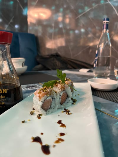 The image, taken in Basiglio, Italy, presents a close-up view of a dining table, likely in a restaurant. The central subject is a white rectangular plate holding three pieces of sushi. These appear to be maki or uramaki rolls, featuring white rice encasing a filling of light-pink fish, consistent with salmon. The sushi rolls are garnished with finely chopped green herbs, resembling parsley or cilantro, and scattered small, light-green to brown fragments, possibly crushed pistachios or another spice. Dark brown sauce splatters, indicative of soy sauce or a similar condiment, are irregularly spread across the pristine white plate.

In the lower foreground, a clear glass bottle with a distinctive red cap contains a dark liquid, identified by its label as soy sauce. Visible text on the label includes "KAN" and "TRADICIONAL ELABORADA," suggesting a traditional brand of soy sauce.

The background features a dark, reflective table surface, possibly marble or a similarly patterned material, with soft, diffused reflections of ambient lighting. Partially visible objects in the background include a tall, clear glass bottle with a blue and white striped label, likely a water bottle, and a small white bowl. A dark, indistinct object, possibly a bag or part of a chair, is also blurred in the distance. The scene depicts a meal being served or about to be enjoyed in an indoor setting, with no people visible. The overall impression is one of a modern and inviting dining atmosphere.