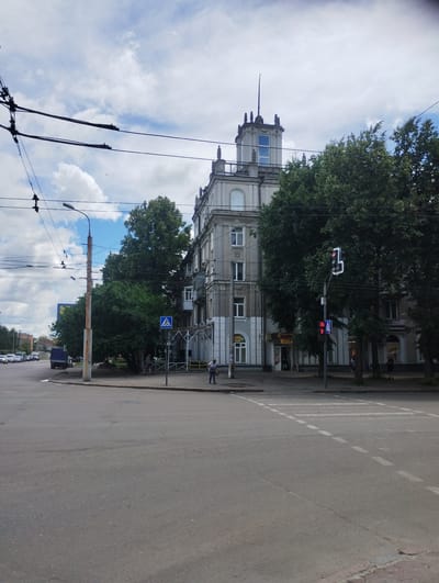 This is a vertically oriented, eye-level, medium shot of a street scene in Sumy, Ukraine. The image captures a building with ornate architectural details, characterized by its light gray facade, arched windows, and decorative molding. A large, mature tree with lush green foliage stands in front of the building, partially obscuring it.
A traffic light, displaying a red signal, is visible hanging over the intersection. Adjacent to it, a pedestrian crossing sign with a blue background and a white walking person symbol is mounted on a pole. The road in the foreground is paved and shows signs of wear, with white dashed lines marking the lanes. Overhead, electrical wires crisscross the frame, suggesting the presence of public transportation or utility lines.
In the background, more buildings and trees can be seen, along with a street with parked cars and a streetlight. A few individuals are present on the sidewalk and near the building. One person, wearing dark clothing, stands near a scaffolding structure, possibly engaged in work. Another person, in a light blue shirt, is visible further down the street.
The sky is partly cloudy with white cumulus clouds against a bright blue background, indicating it is daytime, likely mid-morning or afternoon. The overall impression is that of a typical urban environment on a clear day. No specific event or emotion is conveyed, and no discernible text is visible in the image.