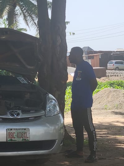 A man in a blue t-shirt and black sweatpants with white stripes stands by an open-hooded silver car. He is facing away from the camera, looking towards the right. The car's license plate reads "RBC-408XL" and displays "ABUJA CENTRE OF UNITY". Behind the man and car is a dirt area with piles of gravel, some green plants, and scattered concrete blocks. In the background, there are simple structures made of corrugated metal and wood, as well as a light-colored minivan. Power lines stretch across the sky. A palm tree trunk is visible to the left of the man and the car. The scene appears to be outdoors, possibly in an informal auto repair or storage area, in Suleja, Nigeria.