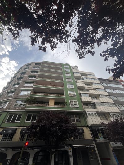 This is a low-angle, wide shot of a curved apartment building with a green facade on the lower floors and a grey and white facade on the upper floors. The building features numerous windows and balconies, with white railings. Dark trees with dense foliage partially obscure the view of the building, with their branches and leaves silhouetted against a bright, cloudy sky. The time of day appears to be daytime, with diffused sunlight.
The lower floors of the building house businesses, indicated by visible signage. Text on the signs includes "ATEMPO EVENTOS," "LIEU," and "Estrella Galicia," suggesting a commercial area. A traffic light showing a red pedestrian signal is visible on the right side of the image, further indicating a street scene. The architecture suggests an urban environment, likely in a European city. No people are visible in the frame, and the overall mood is neutral, focusing on the architectural and natural elements of the scene.