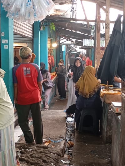 The image shows a narrow street in Kutawaringin, Indonesia, where people are walking, shopping, and talking. The location is a narrow alleyway, with wooden structures and concrete walls. The alleyway is cluttered with loose items. There are six people in the image, all wearing typical Indonesian clothing. One person is wearing a red shirt and appears to be talking to someone. Another person is wearing a yellow headscarf, sitting on a stool, and looking to the right. A third person is wearing a blue headscarf and holding a bag. A fourth person is wearing a black headscarf and is looking at the camera. A fifth person is wearing a white headscarf and is walking past. A sixth person is wearing a blue shirt and is walking toward the camera. The image was captured in a brightly lit environment, which seems to be during the day. There is a dark, black umbrella on the right side of the image, and a bag on the floor to the left of the image. The alleyway is dirty, with dirt and debris scattered on the ground. The scene is a casual and informal setting, and the people appear to be engaged in everyday activities.