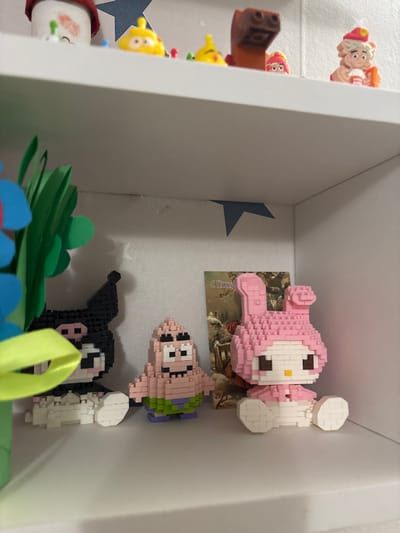 The image displays an interior scene, likely within a residence in Krasny Klyuch, Russia, featuring two white shelves mounted against a light-colored wall.
On the upper shelf, visible from below and partially obscured, are several small, colorful figures. From left to right, these include a white object with red markings and green protrusions, two yellow figures with small red pegs, a brown block-like figure, a small pink/red character, and a beige character wearing a red hat and holding a white object.
The lower shelf, occupying the majority of the frame, exhibits three figures constructed from small building blocks, arranged from left to right. The leftmost figure is a black and white Kuromi character, seated, with a yellow-green ribbon partially obscuring its left side. Adjacent to it is a standing, light pink Patrick Star character, wearing green shorts with purple trim. To the right, a seated pink My Melody character is visible, featuring a pink hood with prominent ears and a white face and body. Behind the Patrick Star figure, a small rectangular card with the Cyrillic text "С Новым" (Happy New) is visible, depicting a snowy, rustic scene with trees. To the far left of the lower shelf, a blurred arrangement of green paper cutouts resembling leaves or grass, accompanied by a blue flower-like shape and a prominent yellow-green ribbon, partially obscures the Kuromi figure and extends upwards.
The walls are light-colored, possibly white or off-white, with several irregular, dark blue star-shaped cutouts or stickers affixed to them, particularly in the corners above the lower shelf and behind the upper shelf. The shelving unit itself is entirely white. Lighting appears even and artificial, typical of an indoor environment. No individuals are present in the scene.