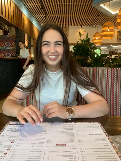 A young woman with long, dark hair is smiling at the camera, sitting at a table in a restaurant. She is wearing a white, textured top and a gold-colored watch on her left wrist. In front of her is a menu with various food and drink items listed in Russian. The restaurant has a rustic interior with wooden beams on the ceiling and decorative woven panels on the wall. In the background, another woman is visible, and there are plants and light fixtures throughout the space. The scene suggests a casual dining experience. The image does not provide specific clues to pinpoint the location within Novorossiysk, Russia.