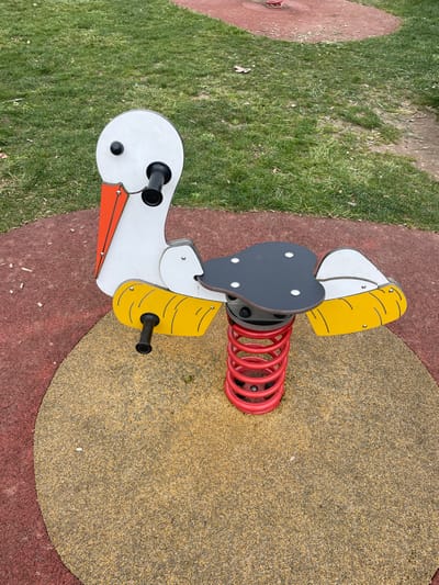 A children's spring rider, shaped like a white stork with an orange beak, black eyes, and yellow wings outlined in black, is centrally positioned on a playground in Holtzheim, France. The rider features a black seat, two black cylindrical handles near the head, and two black cylindrical footrests extending from the yellow wing sections. It is supported by a red coiled spring anchored into a circular yellow and beige granular safety surface. This circular surface is surrounded by a larger area of red granular safety surfacing. Beyond the safety surfacing, the environment transitions to green grass with sparse brown patches and scattered organic debris. A dirt path is visible in the upper right background, and a faint red structure, possibly another playground element, is partially visible further in the background. No individuals are present in the scene, and no actions or interactions are depicted.