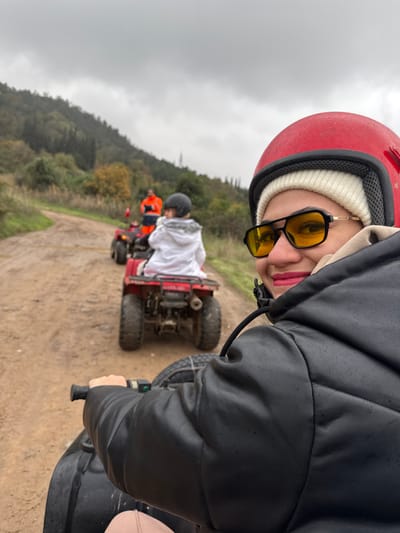 The image depicts a group of three individuals riding ATVs on a muddy dirt path in a rural, hilly area within Çiftlikköy, Turkey, under an overcast sky. In the immediate foreground, the right side of an individual is visible, wearing a red helmet over a white beanie, black-framed sunglasses with yellow lenses, a black padded jacket, and holding the black handlebar of an ATV. This person has pink lipstick and is smiling, looking towards the viewer. Directly in front, a second person, appearing to be a child, rides a red, mud-splattered ATV, wearing a black helmet and a white patterned jacket. Further along the winding dirt path, a third person on another ATV is visible, wearing an orange jacket or vest. The environment consists of green, tree-covered hills in the background and sparse vegetation flanking the unpaved path.