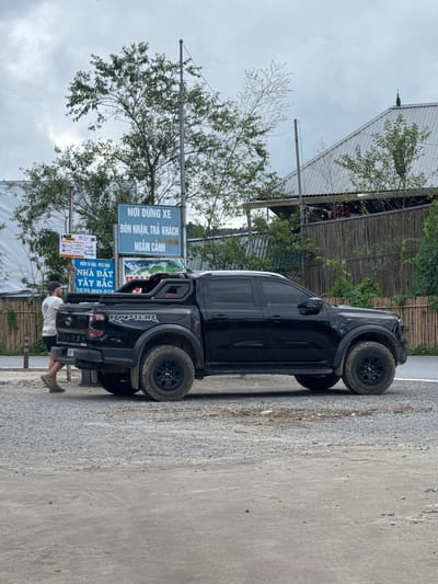This image captures a man standing beside a black pickup truck in Tu Le, Van Chan District, Yen Bai Province, Vietnam, specifically near a location designated for parking, passenger drop-off/pick-up, and sightseeing, as indicated by the blue sign reading "NƠI DỪNG XE ĐÓN NHẬN, TRẢ KHÁCH NGẮM CẢNH."
The man, with short dark hair and a baseball cap, wears a white t-shirt, dark shorts, and athletic shoes. He is positioned on the left side of the truck's rear, facing away from the camera, appearing to interact with or stand near the vehicle.
The vehicle is a black Ford Ranger Raptor pickup truck, identifiable by the "RAPTOR" branding on the bed and "RANGER" on the tailgate. It features a black roll bar or sport bar in the truck bed with "BUFFALO" written on it. The truck is equipped with aggressive off-road tires and black rims, and its body appears somewhat dirty, suggesting recent travel on unpaved surfaces. The rear license plate is partially obscured but shows "29K".
Behind the man and the truck, two informational signs are visible. The prominent blue sign is for the parking and passenger area. Below it, a smaller sign advertises "NHÀ ĐẤT TÂY BẮC" (Northwest Real Estate) with Vietnamese phone numbers. Further to the left, a white sign with various smaller labels, possibly directions or regulations, is partially visible.
The ground surface in the foreground and beneath the truck is a mix of gravel and packed dirt/concrete. In the background, there are numerous trees with green foliage and utility poles. A structure with a corrugated metal roof and a bamboo-slat fence is visible behind the trees and the truck, suggesting a building or a viewing platform. The sky is overcast and grey, indicating a cloudy day.