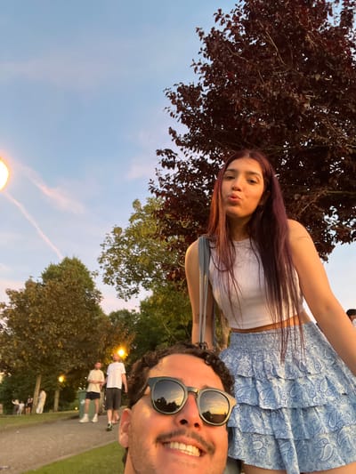 This is a selfie taken outdoors, likely in a park in Vila Nova de Gaia, Portugal, as suggested by the presence of trees, pathways, and people engaged in leisure activities. The sky is a gradient of pale blue with wispy pink clouds, indicative of either dawn or dusk. A large, dark-leafed tree dominates the upper right portion of the frame.
In the foreground, the lower half of a man's face is visible, wearing sunglasses that reflect the sunset sky. He has curly dark hair and a mustache, and he is smiling. Behind him and slightly to the left, a woman is positioned. She has long, dark hair with purple undertones and is making a kissing face. She is wearing a white crop top and a blue, patterned ruffled skirt. She also has a grey bag slung over her shoulder.
In the background, several people are visible walking along a pathway. Two men are walking side-by-side, one in a light-colored t-shirt and shorts, the other in a white t-shirt and dark shorts. Other figures are more distant and less distinct. Several lampposts are scattered throughout the background, one of which is prominently lit on the left side of the image. The ground appears to be a mix of grass and paved paths.