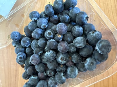 A close-up, top-down view shows a clear plastic container filled with a pile of fresh blueberries. The blueberries are mostly dark blue with a slight dusty bloom on their surfaces. Some show small stems or remnants of them. The container is positioned on a light brown wooden surface. The background shows a light blue, out-of-focus object, possibly a table or a wall. There are no people or animals present in the image. The scene appears to be indoors, with the lighting suggesting natural light from a window or overhead source. The image is in focus and shows a high level of detail on the blueberries.