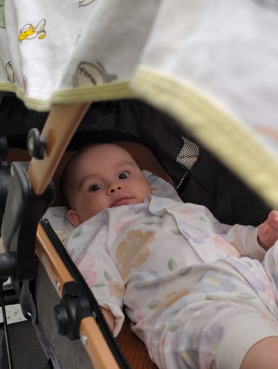 A baby lies in a stroller, face up, with the camera pointing down into the stroller. The baby wears a white floral patterned long-sleeved outfit. The stroller has a tan and yellow trim and a white canopy with a pattern of yellow bananas and baseballs. The location cannot be determined from the image.