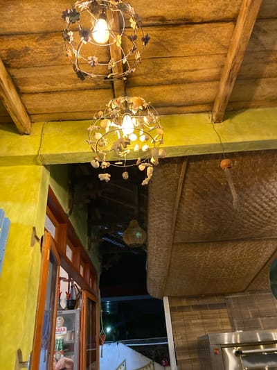 The image depicts an interior shot of a restaurant or cafe, possibly in Karimunjawa, Indonesia, with a focus on decorative elements and architectural features. The scene is illuminated by ambient lighting, casting a warm glow and suggesting it is evening or nighttime.
Dominating the foreground are two ornate spherical pendant lights, each with a delicate metal frame adorned with floral and shell-like embellishments. One light, to the left, is closer to the viewer and reveals a bright bulb within, illuminating the surrounding area. The other light, to the right, is slightly further away and also emits a warm light. These lights hang from a wooden ceiling constructed of vertical planks.
The walls are painted a vibrant, textured yellow-green, providing a striking contrast to the natural wood of the ceiling and accents. A prominent wooden beam runs horizontally across the upper left portion of the image, adding to the rustic aesthetic. Above the wooden ceiling, a woven bamboo material forms a textured canopy, suggesting a tropical or island-inspired design.
In the mid-ground, more architectural elements are visible, including wooden shelving or display units. These structures have glass fronts, revealing glimpses of items stored within, though they are not clearly identifiable. One of these units appears to be a small refrigerated display case, with bottles and packages visible inside. To the far right, a stainless steel appliance, possibly a commercial kitchen fixture, is partially visible.
A small, decorative clay pot hangs from a string in the upper right section of the image, adding another handcrafted element to the decor. The overall impression is one of a cozy, atmospheric establishment with a strong emphasis on natural materials and artisanal touches, likely catering to a relaxed clientele. The warm lighting and textured surfaces create a welcoming and intimate ambiance.
